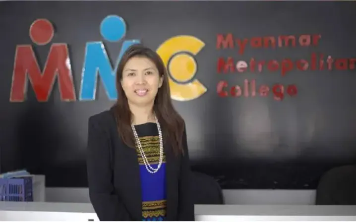 Daw Khin Hnin Soe standing in front of the Myanmar Metropolitan College logo