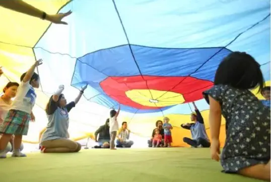 Children and parents playing under a large colourful parachute in a Gymboree play session